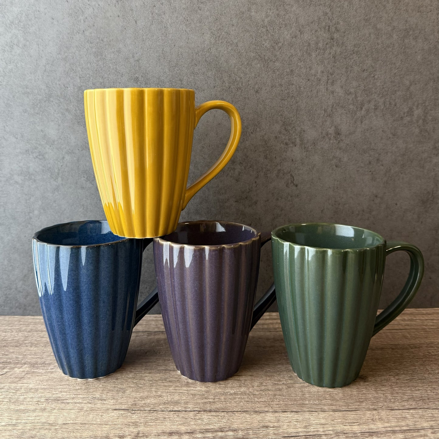 Four colorful mugs (yellow, blue, purple, green) on a wooden surface with a gray background