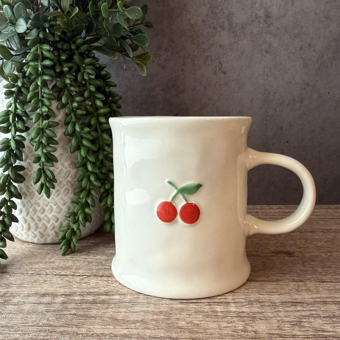 An off-white ceramic mug with a small red cherry and green leaf design on the front, sitting on a wooden surface next to a potted trailing green plant.