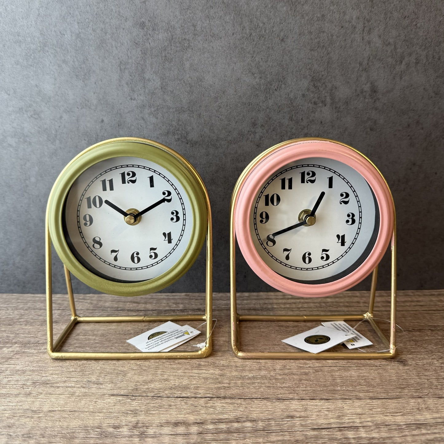 Two small table clocks, one gold and one pink, on a wooden surface with a gray background.