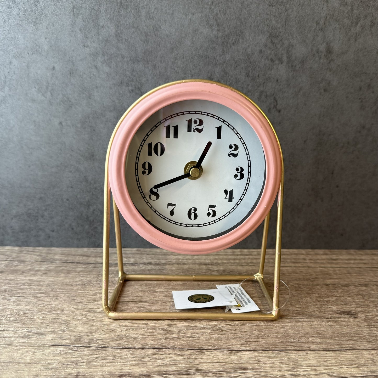 Small clock with pink frame and white face on a wooden surface with gray background