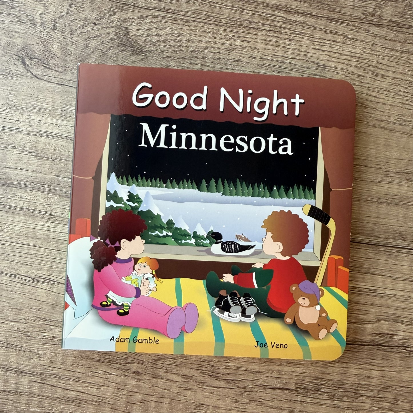 A children's board book titled 'Good Night Minnesota' placed on a wooden surface. The cover shows an illustrated winter scene with two children sitting by a window, a duck by the water, and snowy mountains outside at night.