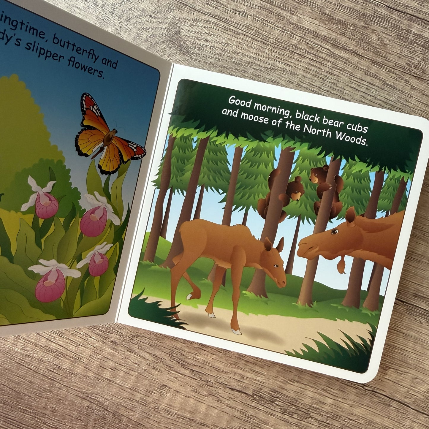 An open children's picture book lying on a wooden surface, showing illustrated pages with text and scenes of a butterfly, flowers, and black bear cubs in a forest.