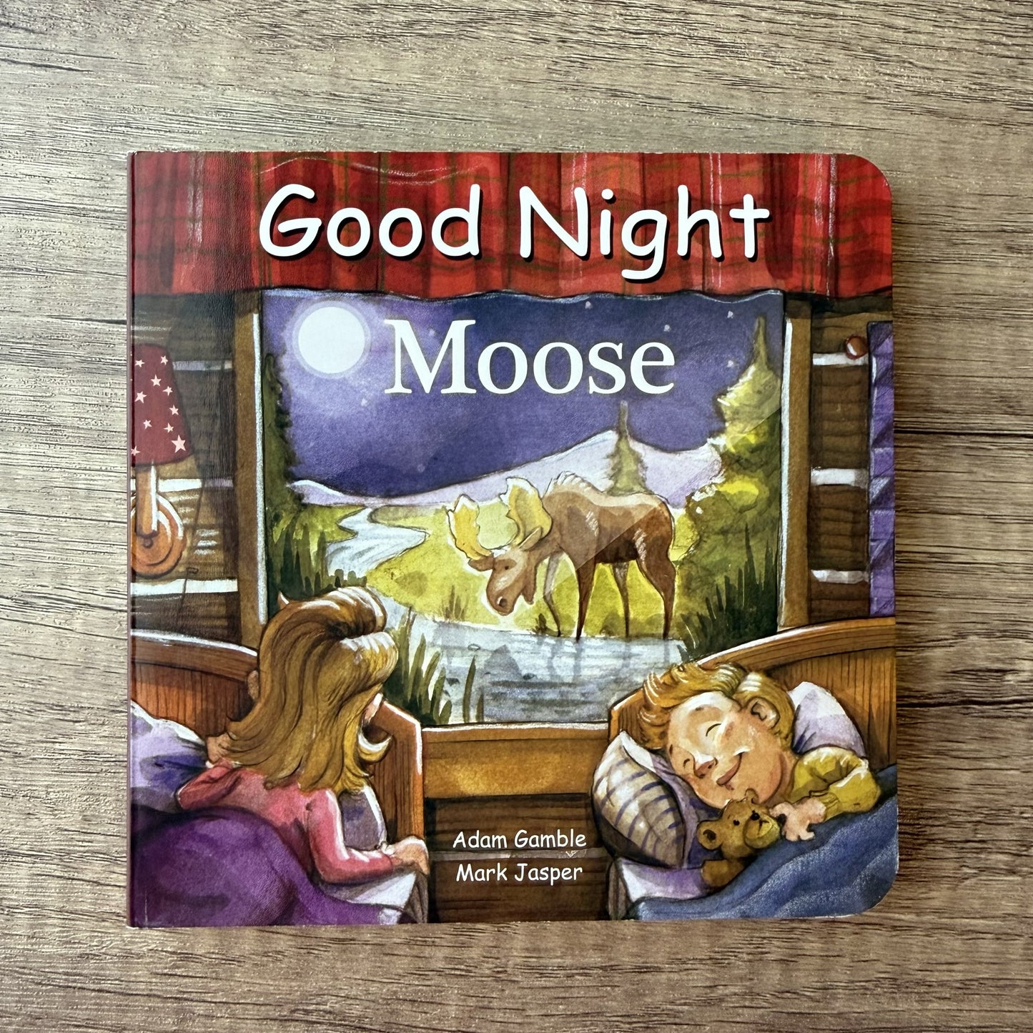 A children's board book titled 'Good Night Moose' by Adam Gamble and Mark Jasper, featuring an illustrated cover of two children in bed looking at a moose outside a window, placed on a wooden surface.