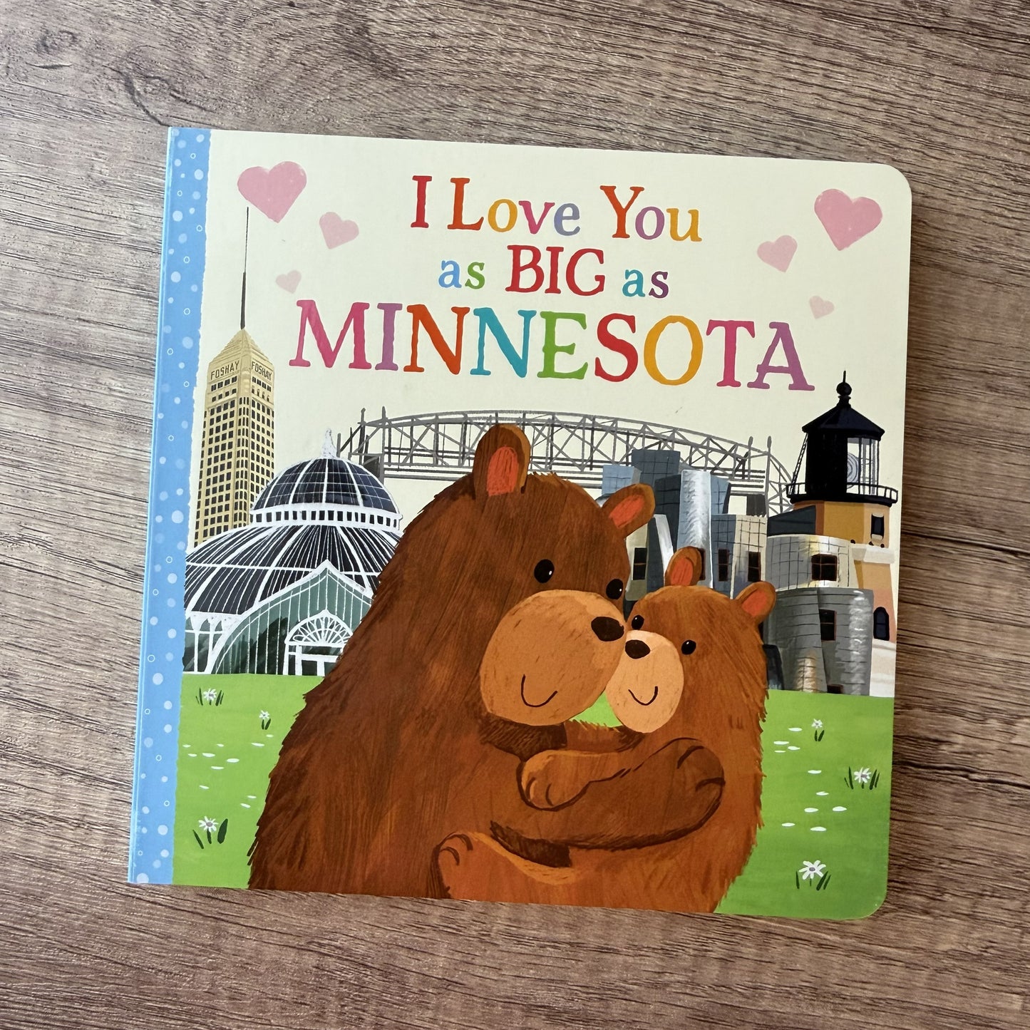 A children's board book titled 'I Love You as BIG as MINNESOTA' lies on a light brown wood surface. The cover shows two brown bears hugging in front of a Minnesota skyline, with pink hearts and a blue dotted border on the left.