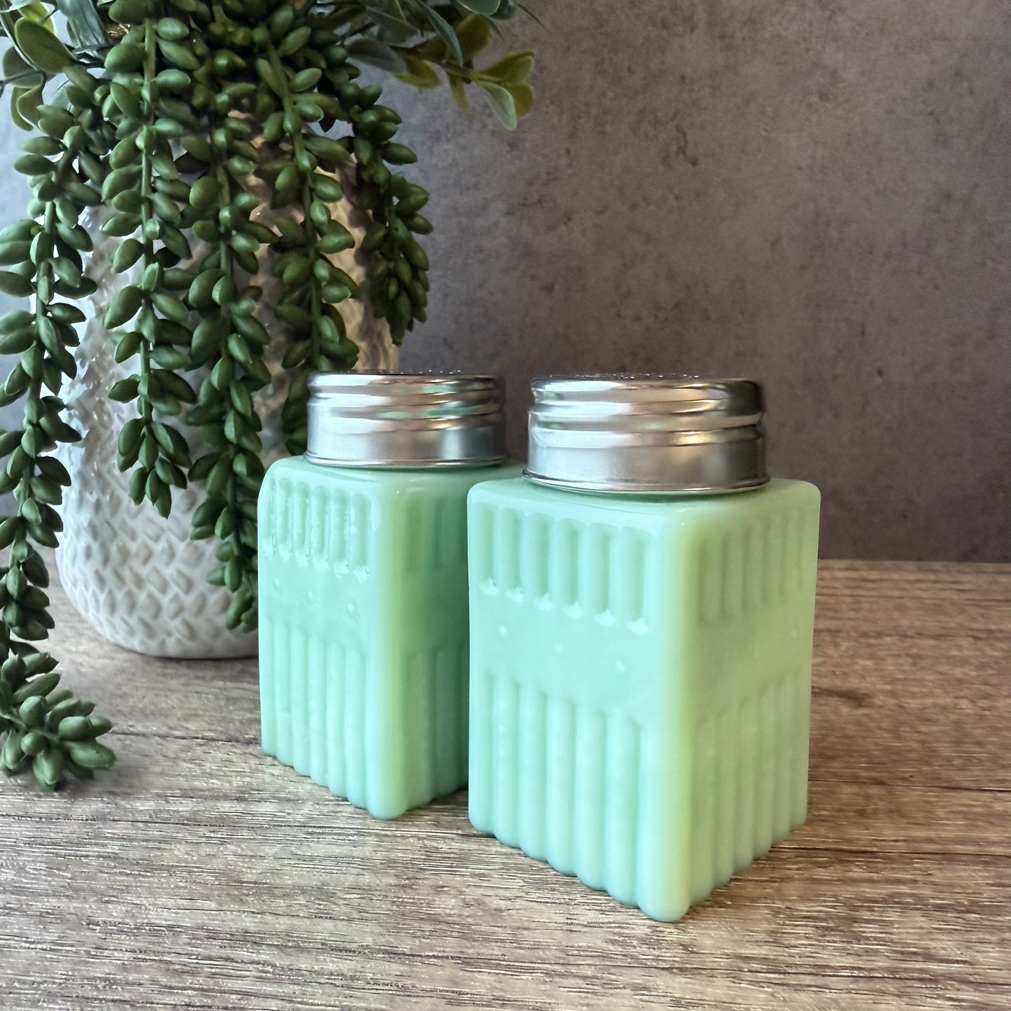 A pair of vintage mint green glass salt and pepper shakers with ribbed sides and silver metal caps, set on a wooden tabletop next to a potted trailing green plant.