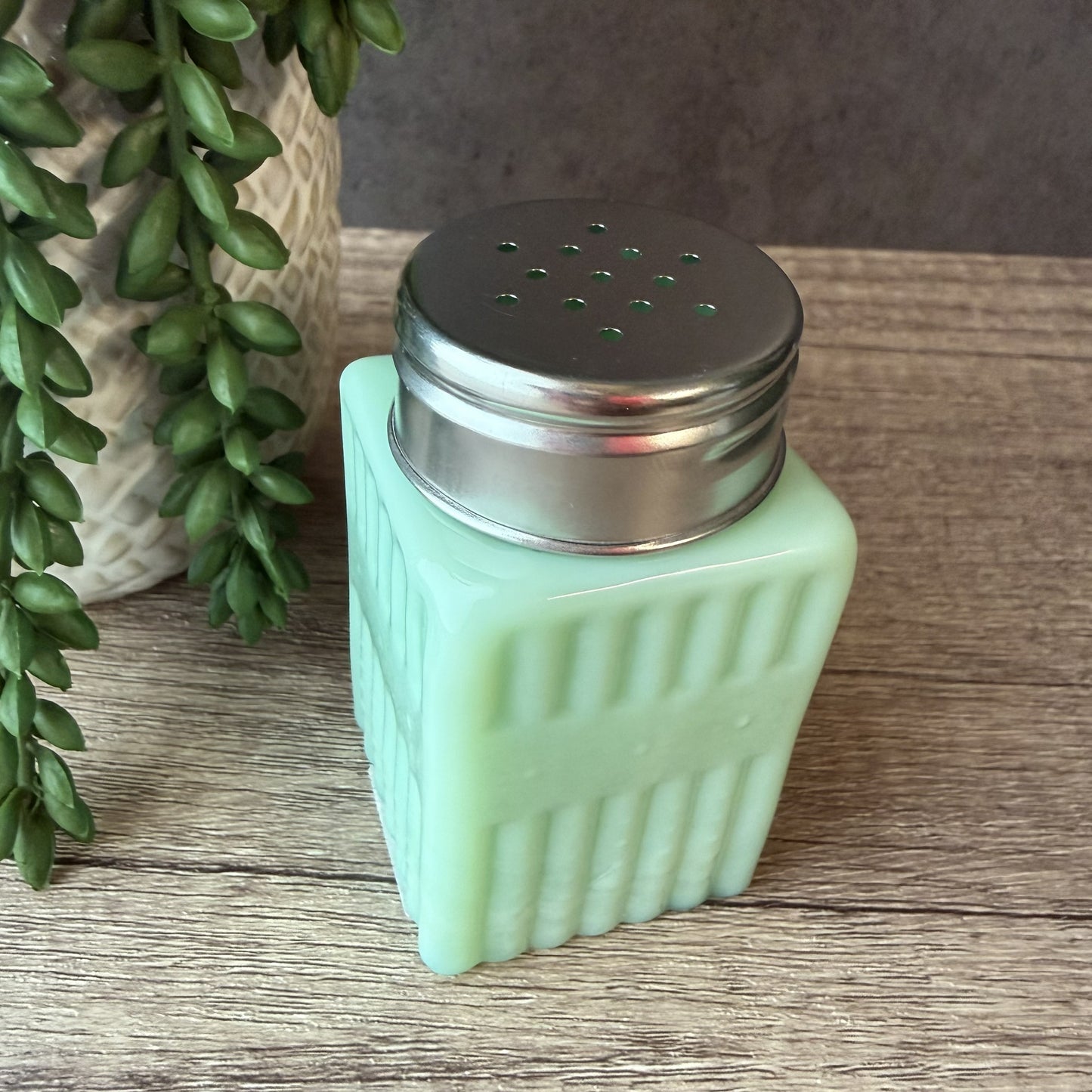 A mint green glass salt shaker with a ribbed square body and a silver metal shaker top, shown on a wooden tabletop next to a potted green plant.