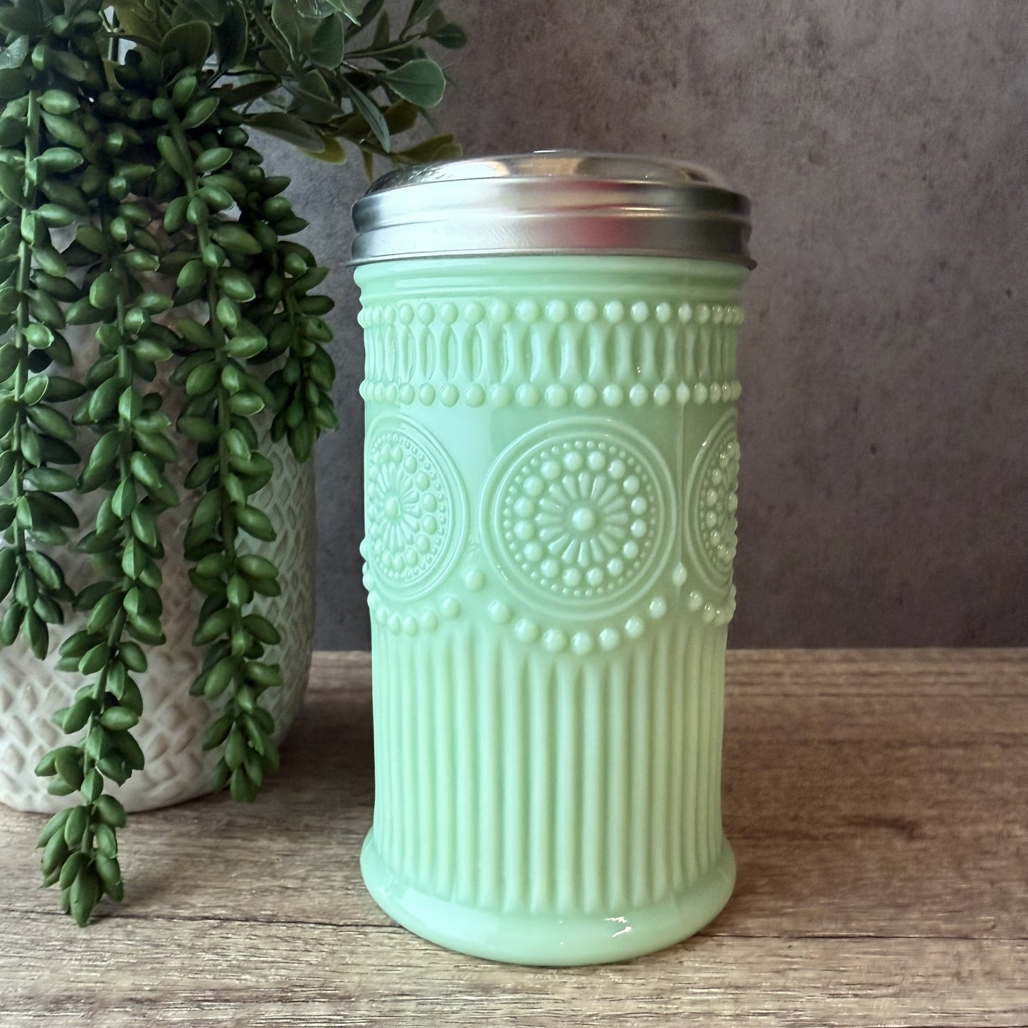 A mint green glass canister with a silver metal lid, featuring embossed circular floral patterns and vertical fluting, displayed on a wooden surface next to a potted green plant.