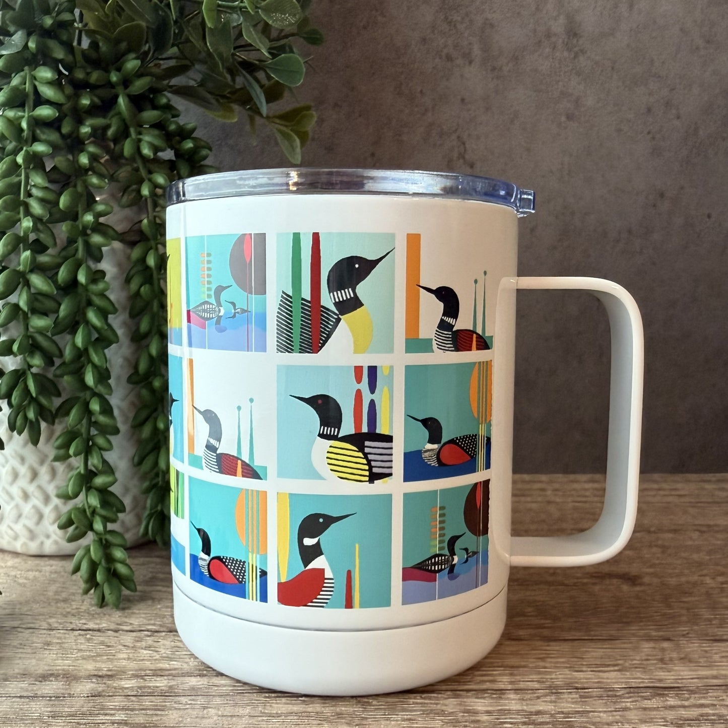 A white insulated travel mug with a clear lid, featuring a colorful grid pattern of loon birds on the front. The mug is set on a wooden surface next to a potted green plant against a textured gray wall.