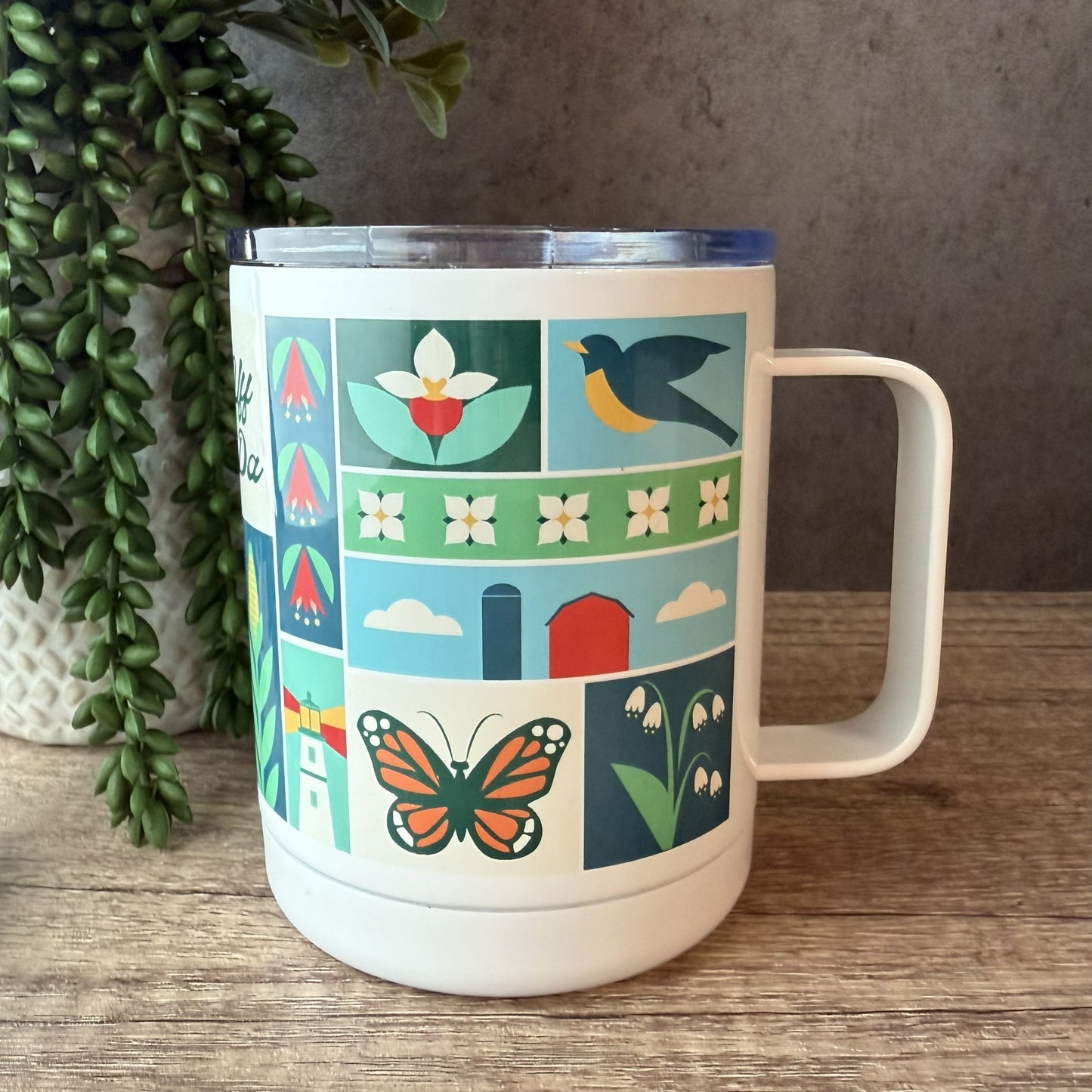 A white insulated travel mug with a colorful spring-themed pattern of flowers, birds, barns, and a butterfly, sitting on a wooden table next to a potted plant.