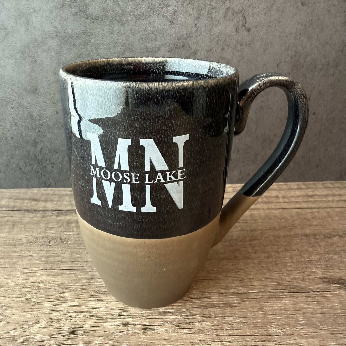 A ceramic coffee mug with a glossy black upper half, a matte beige lower half, and a curved handle. The mug features a white printed graphic of a moose silhouette and the text 'MN MOOSE LAKE'.