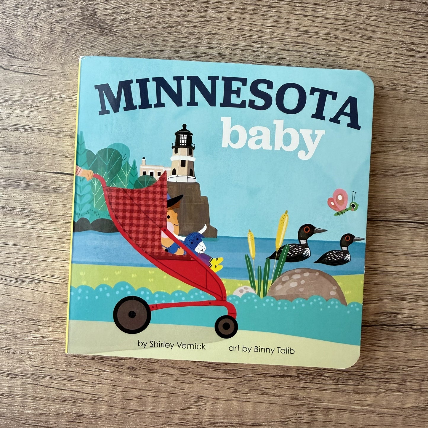 A children's board book titled "MINNESOTA baby" lies on a wooden surface. The cover shows an illustrated scene with a baby in a red stroller near a lighthouse, with ducks, grass, and a butterfly.