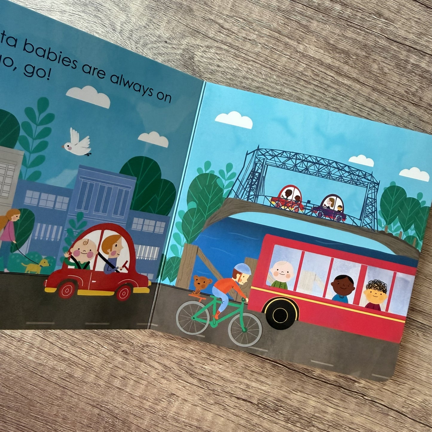 An open children's picture book lying on a wood surface, showing illustrated city scenes with text on the left page and a red bus, cyclist, car, and buildings on the right page.