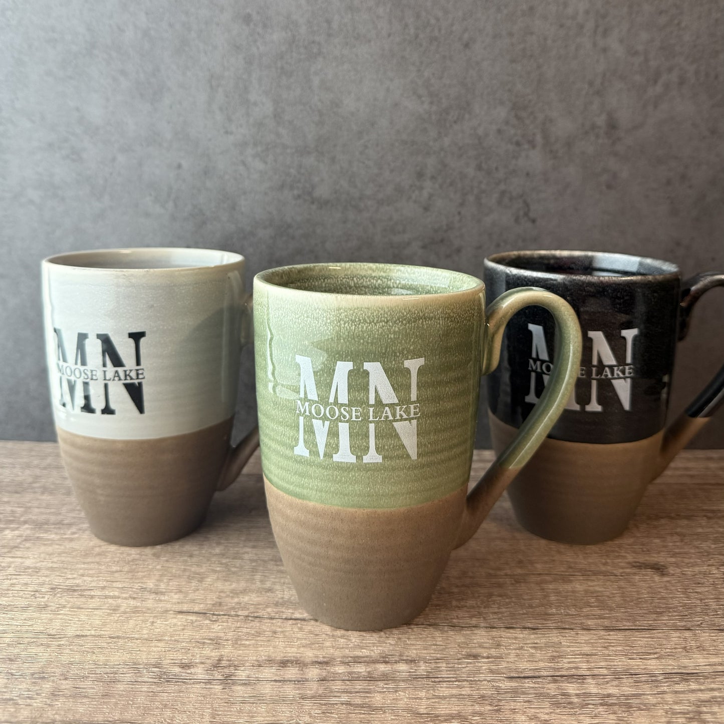 Three ceramic mugs with a two-tone finish, featuring the text 'MN MOOSE LAKE' in white. The center mug is light green and brown, while the mugs on the left and right are beige and brown, and black and brown respectively.