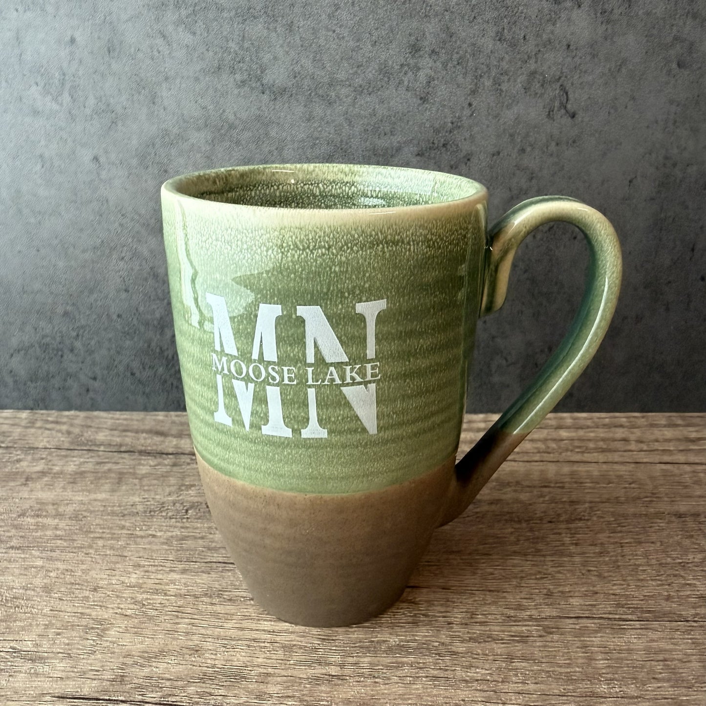 A ceramic mug with a two-tone green and brown glaze, featuring the white text 'M N MOOSE LAKE' on the front, set on a wooden surface against a gray textured background.