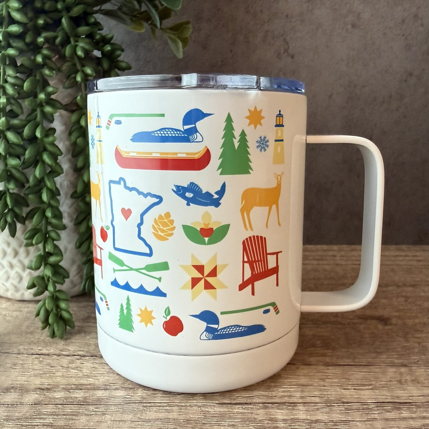 A white insulated travel tumbler with a clear lid and handle, decorated with colorful outdoor-themed illustrations including a loon, trees, a lighthouse, an adirondack chair, a deer, and a maple leaf. The tumbler is photographed on a wooden surface next to a potted green plant.