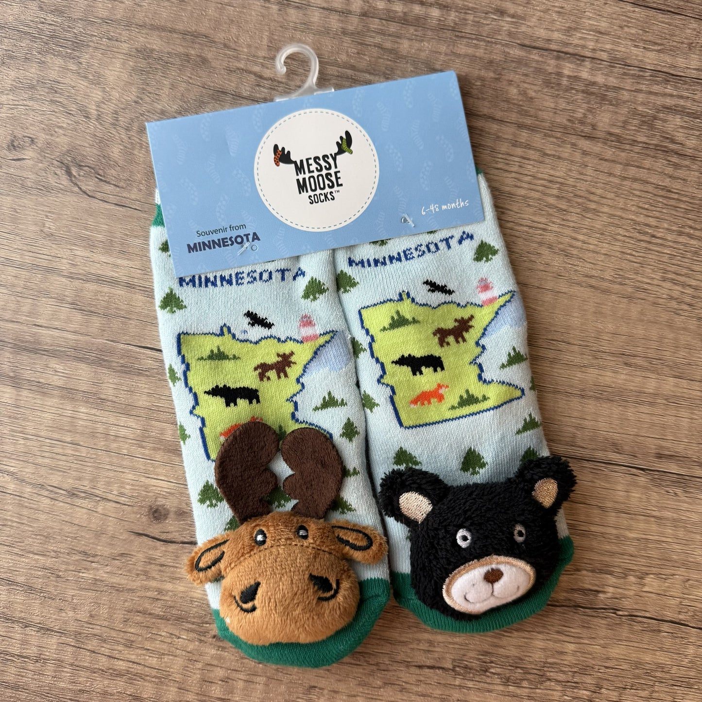 A pair of children's Minnesota-themed socks in their retail packaging, featuring a light blue background with green state maps, woodland animals, and plush moose and bear feet at the bottom. The package card shows the brand 'Nest Mouse Socks'.