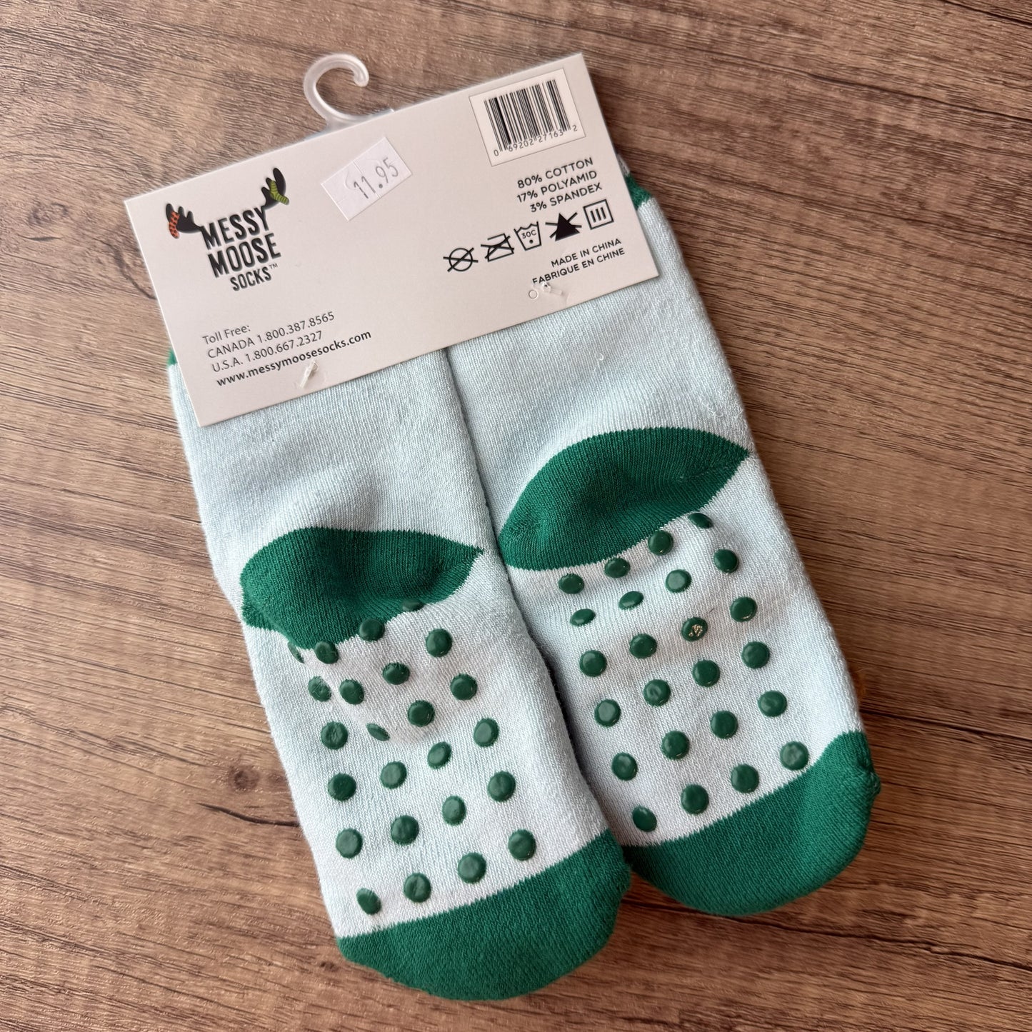 A pair of Messy Moose Socks with green grip dots on a white background, packaged with a cardboard header and shown on a wooden surface.
