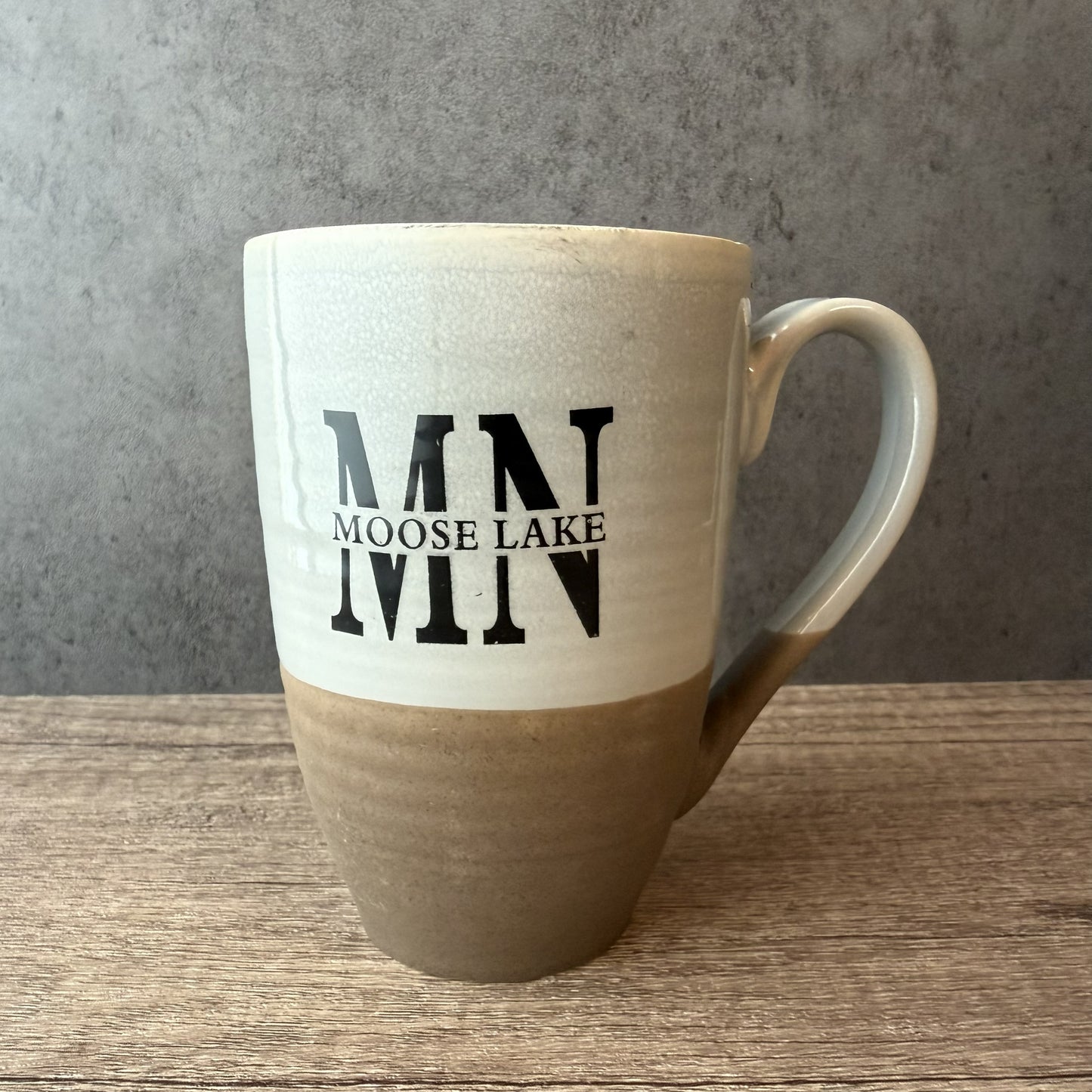 A ceramic mug with a two-tone design, featuring a cream-colored upper half with black 'MN MOOSE LAKE MN' lettering and a brown textured lower half, set on a wooden surface against a gray concrete wall.