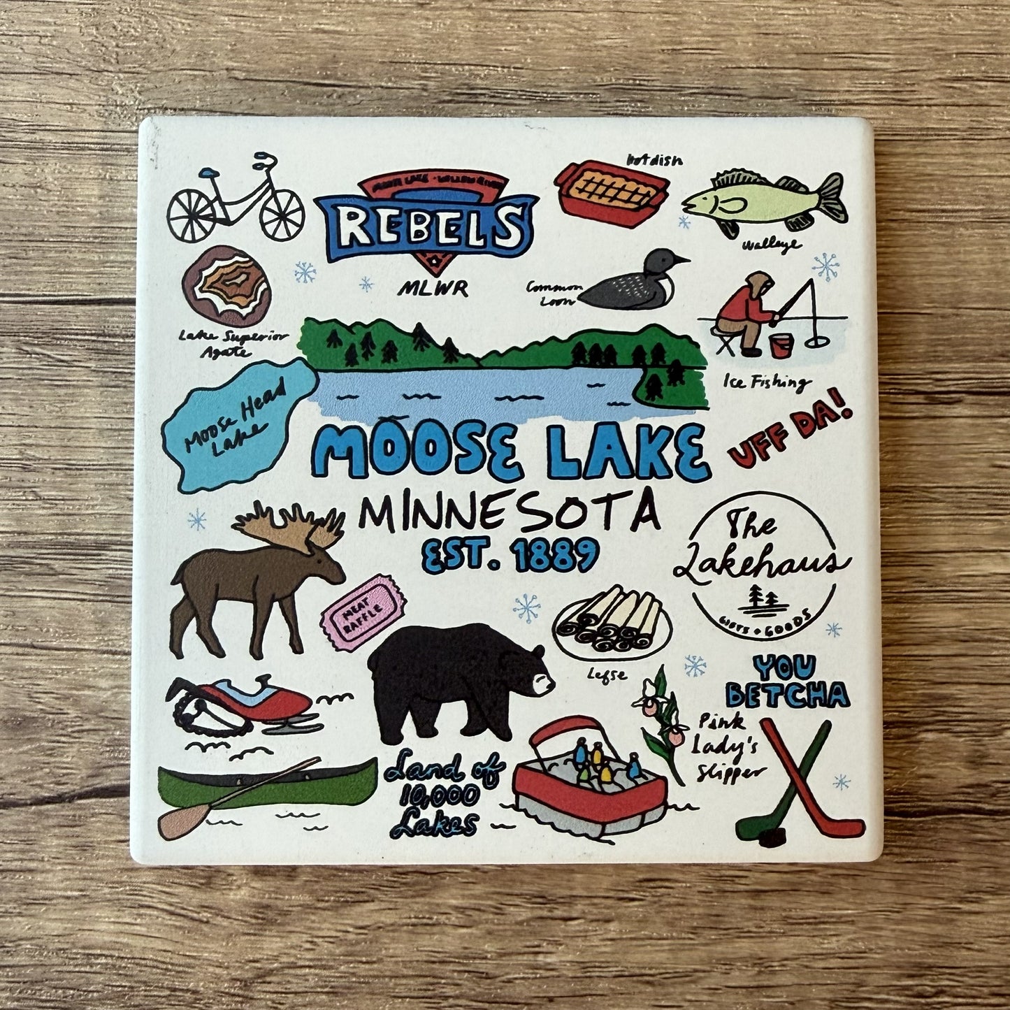 A square ceramic coaster featuring a hand-drawn style illustration of Moose Lake, Minnesota, with various winter and outdoor activities, the word 'Rebels,' and local landmarks, shown on a wooden surface.