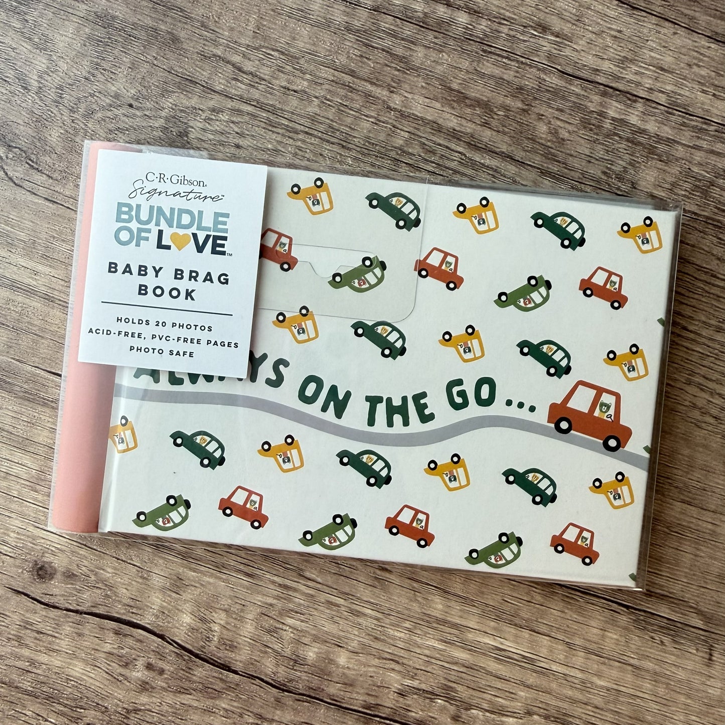 A baby photo album with a pink spine and a clear plastic cover, placed on a rustic wooden surface. The cover has a white background with a pattern of colorful cars and the text 'ALWAYS ON THE GO.
