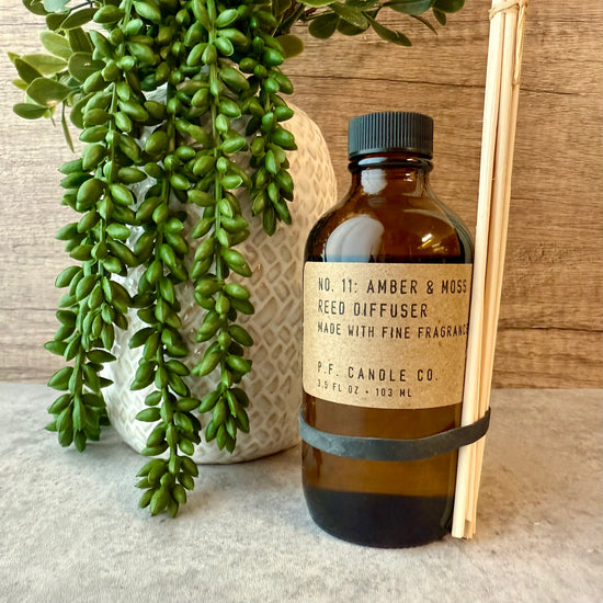 Amber & Moss Reed Diffuser by P.F. Candle Co. with green plant and wooden sticks on a neutral background