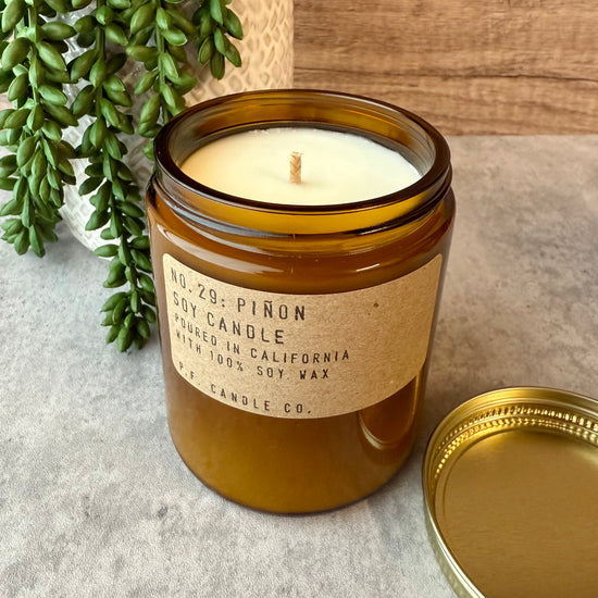Soy candle in a brown glass jar with a label, surrounded by greenery on a textured surface.