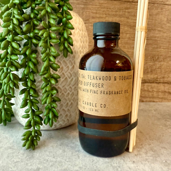 A bottle of teakwood and tobacco-scented diffuser with a label, surrounded by a plant and wooden sticks on a neutral background.