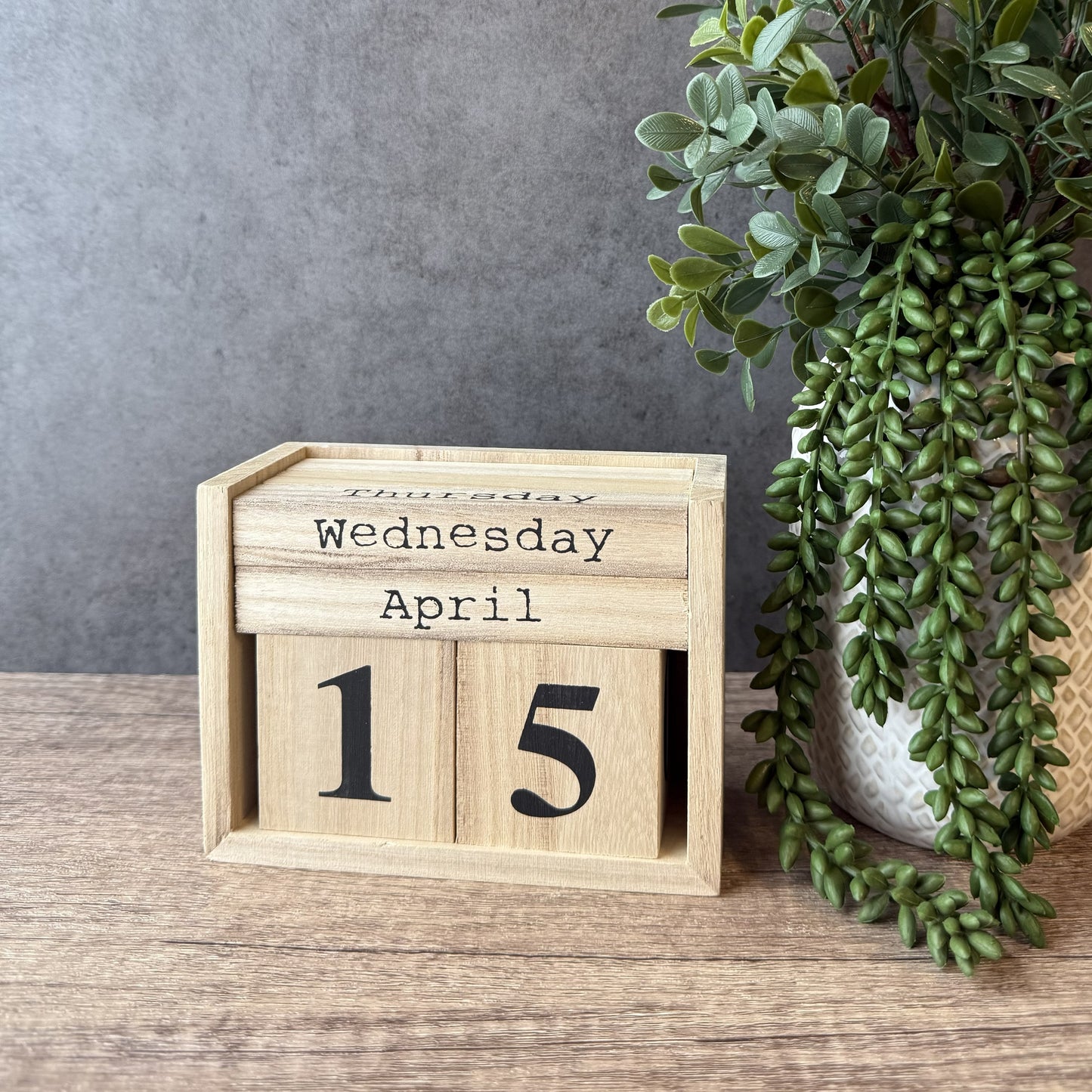 Wooden calendar block showing 'Wednesday, April 15' on a wooden surface with a plant in the background.