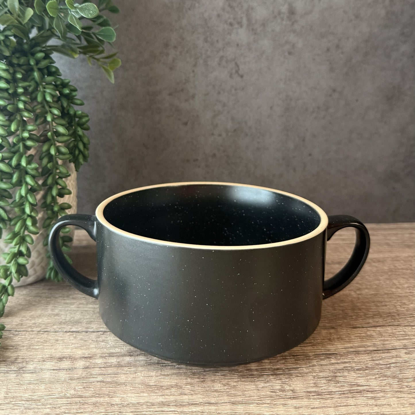 A black speckled ceramic bowl with two side handles and a contrasting beige rim, shown on a wooden tabletop with a potted green plant and a textured gray wall in the background.