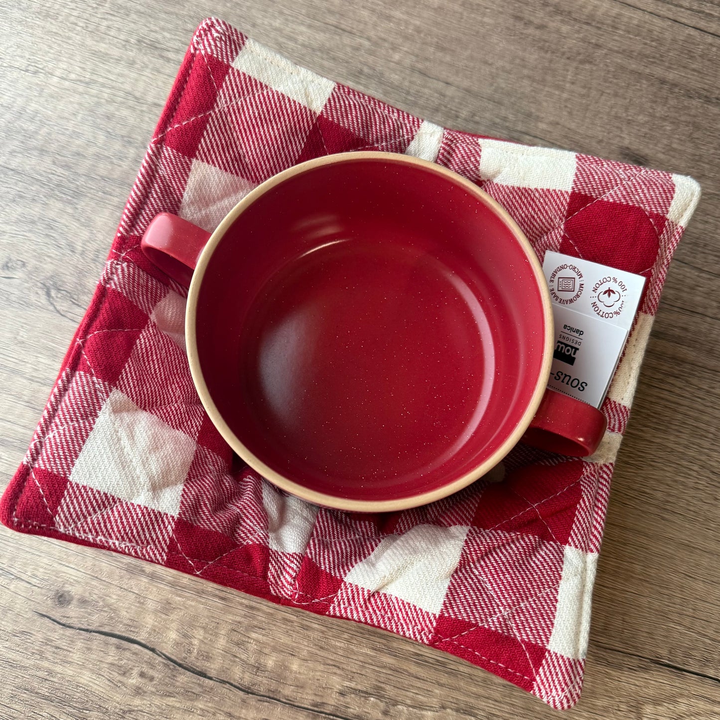 A red ceramic bowl with side handles sits on a red-and-white gingham quilted cloth, placed on a wooden tabletop. A small product tag is attached to the bowl.