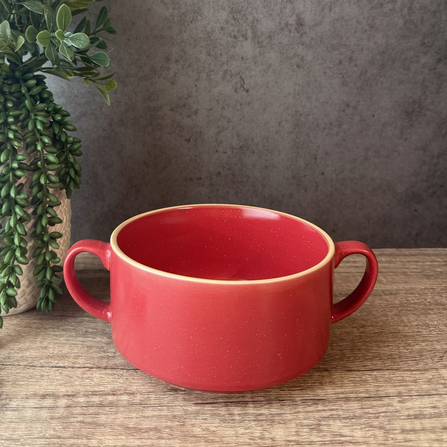 A red ceramic bowl with two side handles sits on a wooden tabletop against a textured gray wall, with a potted green plant partially visible on the left.