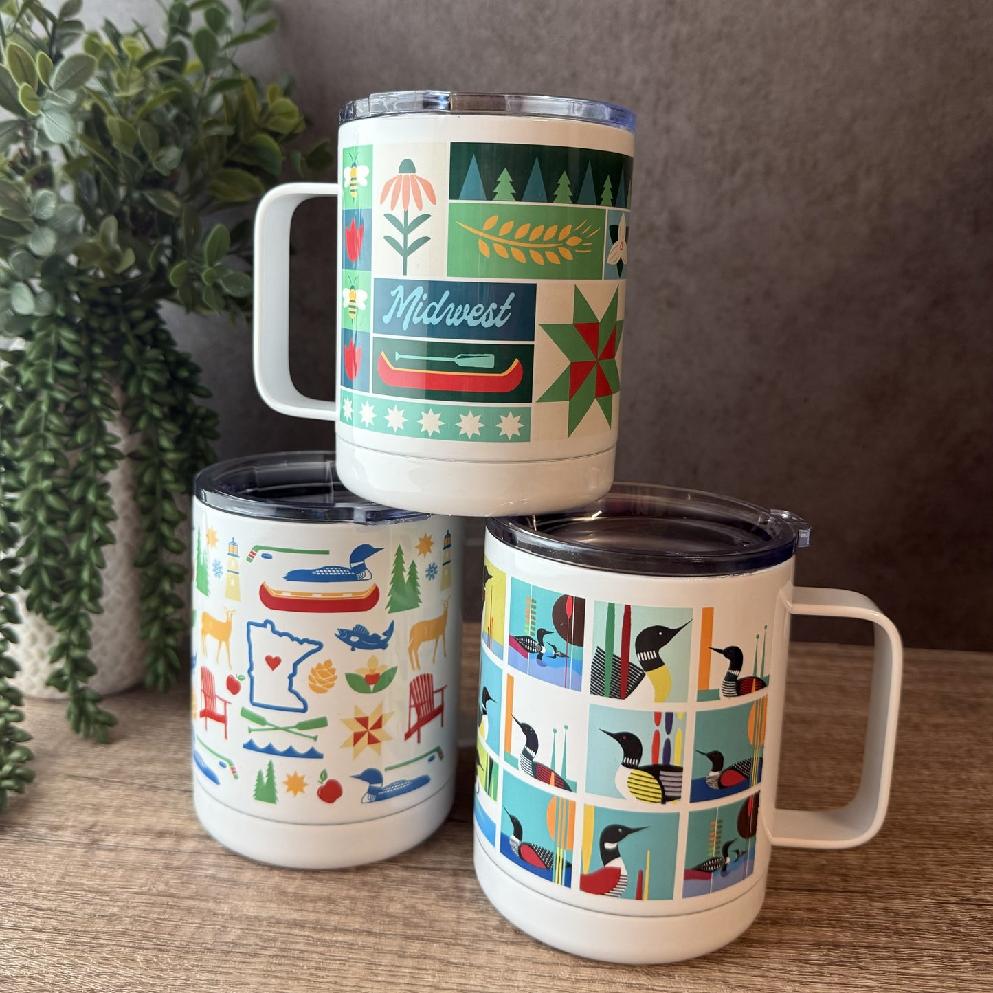 Three insulated stainless steel mugs with lids are stacked on a wooden surface. The top mug is white with a green and red Midwest winter pattern, the bottom left mug is white with multicolor outdoor icons, and the bottom right mug is white with a grid of bird illustrations.