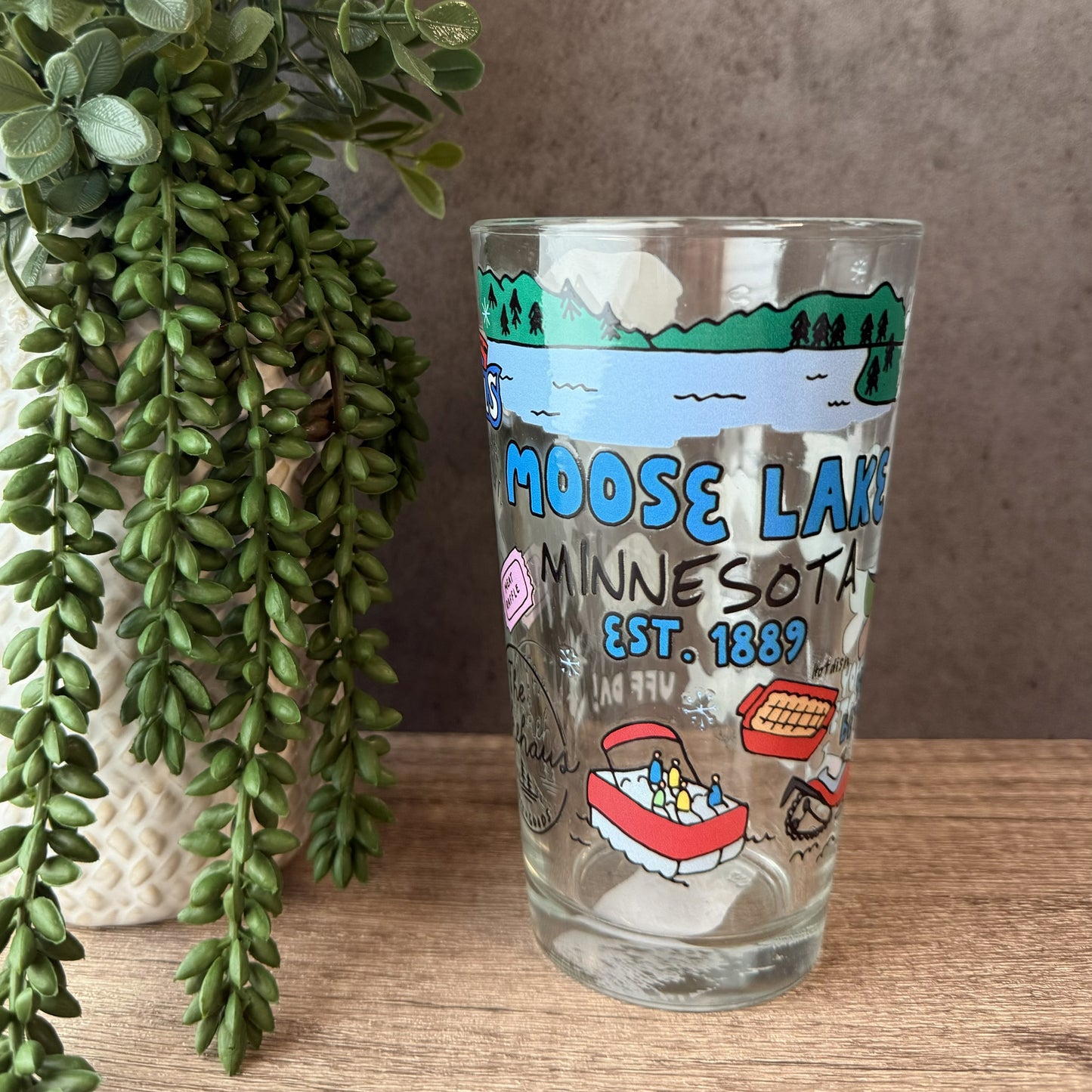 A clear glass drinking tumbler with a colorful Moose Lake Minnesota illustration, set on a wooden surface next to a potted green trailing plant.