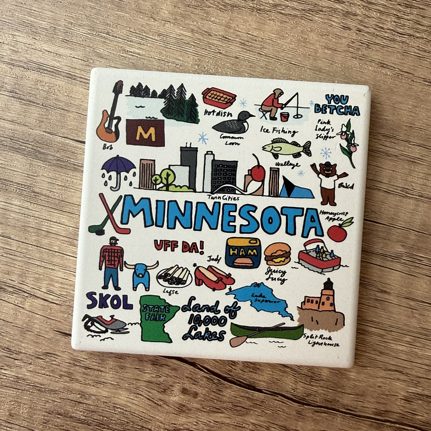 A square white ceramic coaster featuring a colorful hand-drawn Minnesota-themed illustration, placed on a brown wooden surface.