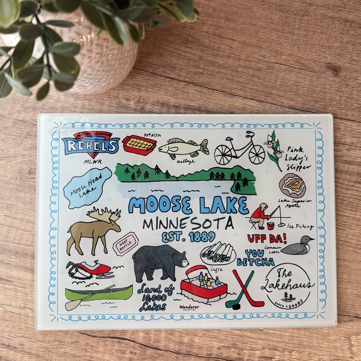 A square illustrated placemat featuring a colorful Moose Lake Minnesota design, displayed flat on a wooden tabletop with a potted plant partially visible in the top left corner.