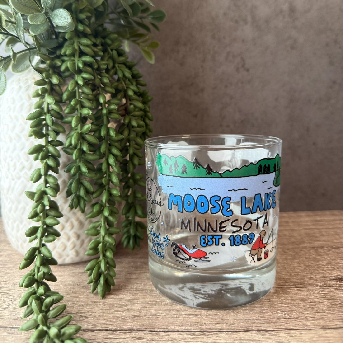 A clear glass tumbler with a colorful 'Moose Lake Minnesota' landscape print, styled on a wooden surface next to a potted trailing green plant.