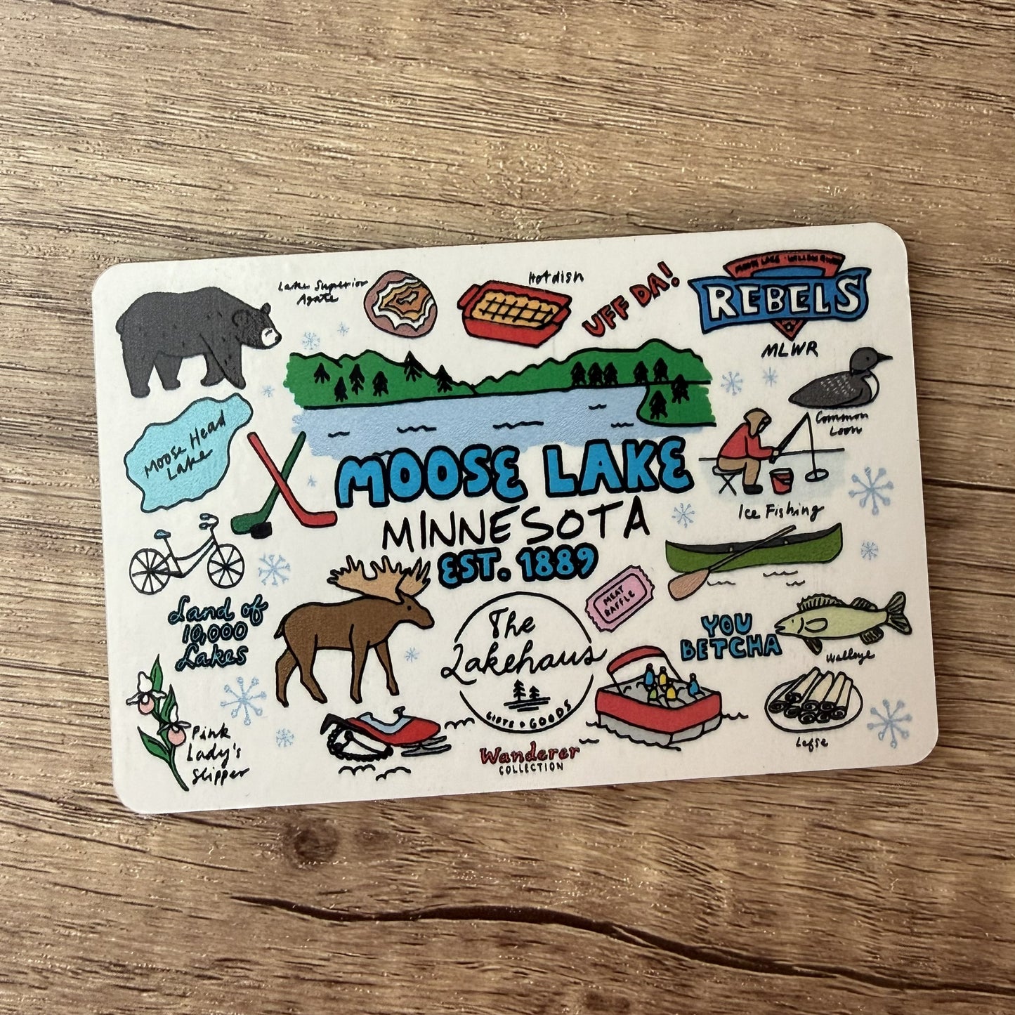 A square white metal sign featuring hand-drawn Moose Lake Minnesota illustrations, including a moose, lake, snowmobile, bicycle, and winter activities, placed on a wooden surface.