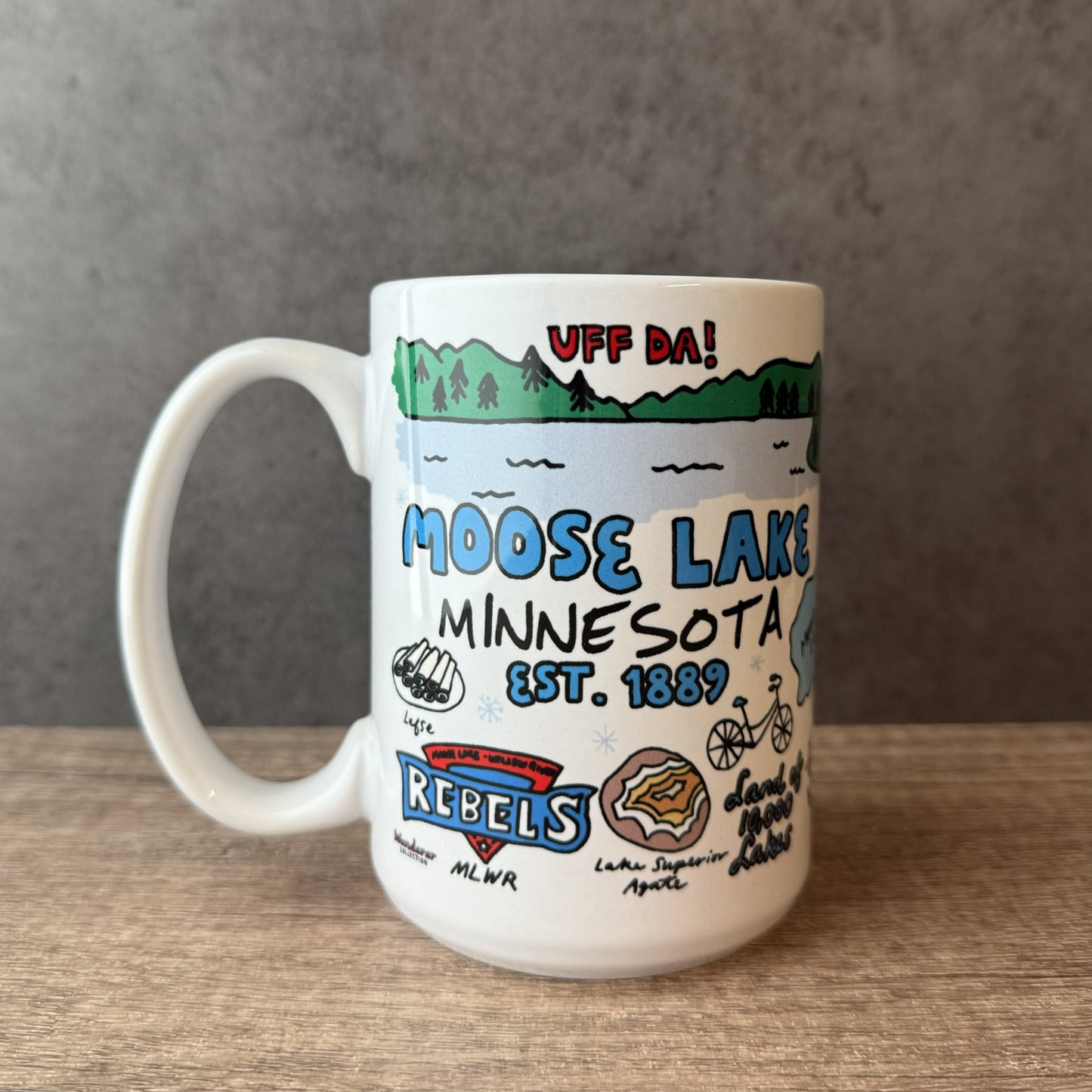 A white ceramic coffee mug featuring a hand-drawn style illustration of Moose Lake, Minnesota, with text reading 'UFF DA!' and 'MOOSE LAKE MINNESOTA EST. 1869', sitting on a wooden surface against a gray background.