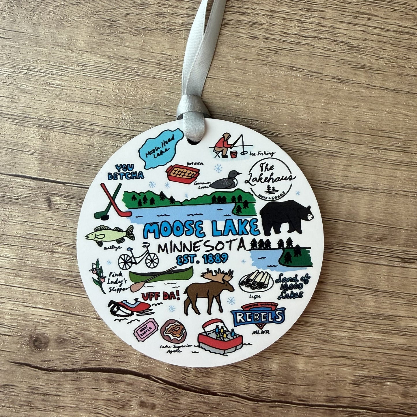 A round white ceramic ornament featuring colorful illustrations and text related to Moose Lake, Minnesota, including a moose, bear, fish, canoe, hockey sticks, and the words 'Moose Lake Minnesota Est. 1939' and 'Reels'.