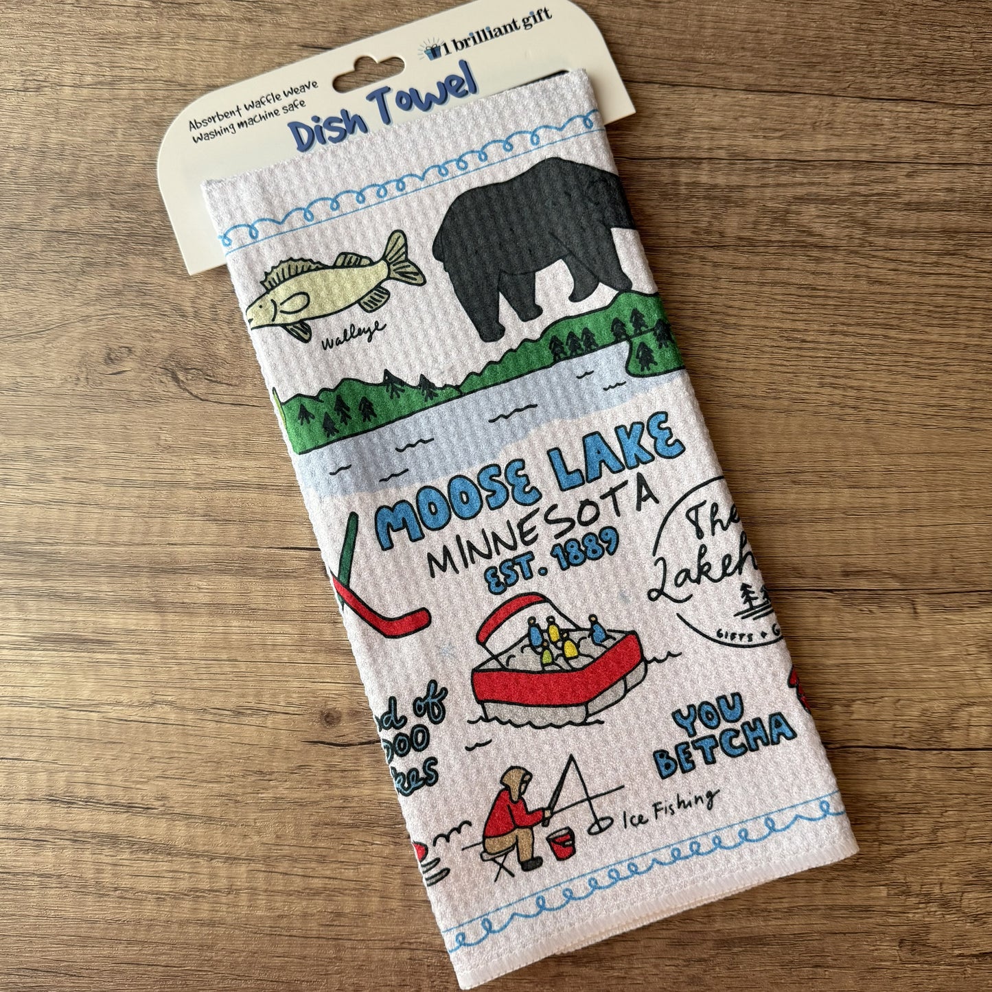 A waffle-weave dish towel with a 'Moose Lake Minnesota' outdoor theme, hanging from its cardboard header on a wooden tabletop. The towel features illustrations of a fish, a bear, a red canoe, and text including 'Moose Lake Minnesota Est.