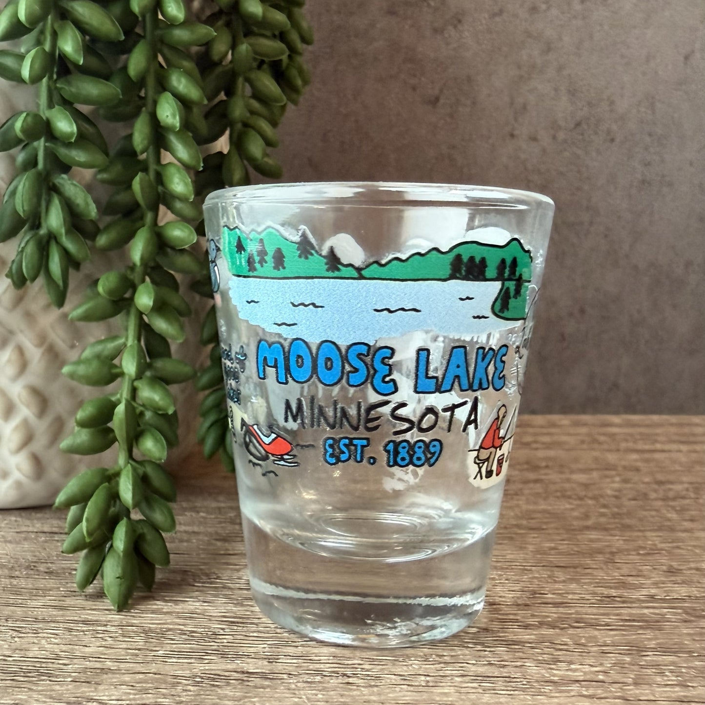 A clear glass shot glass featuring a colorful printed illustration of Moose Lake, Minnesota, with blue text reading 'MOOSE LAKE MINNESOTA EST. 1889', placed on a wooden surface next to a potted green plant.