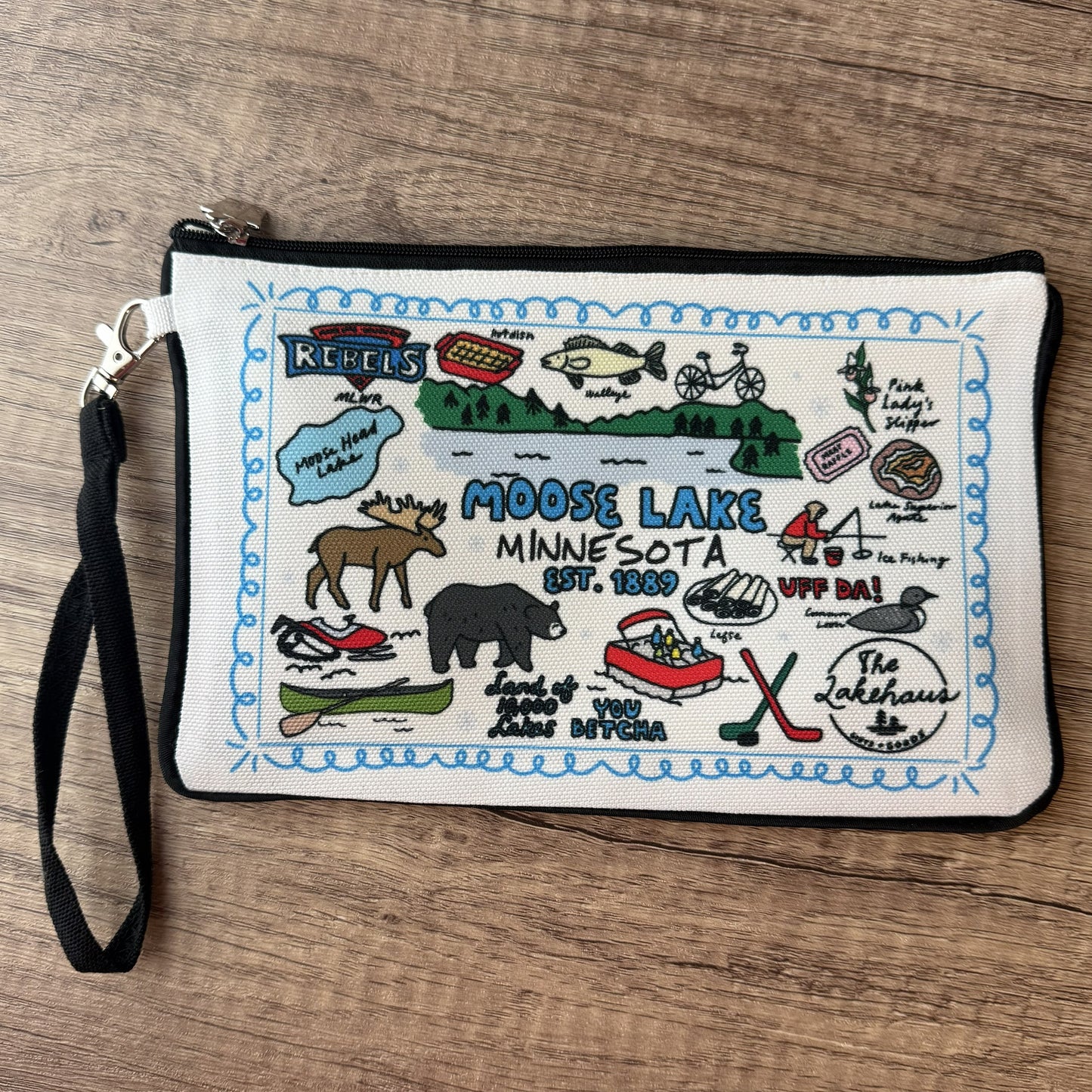 A square canvas clutch pouch featuring a colorful illustrated design of Moose Lake, Minnesota, with a black wrist strap and zipper, placed on a wooden surface.