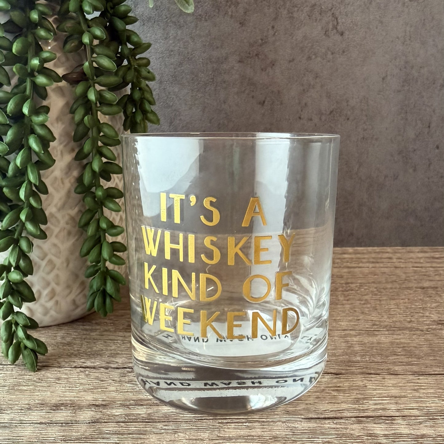 A clear glass rocks tumbler with gold lettering that reads 'IT'S A WHISKEY KIND OF WEEKEND' and 'BINGO MY 21st YEAR', shown on a wooden surface next to a potted trailing green plant.