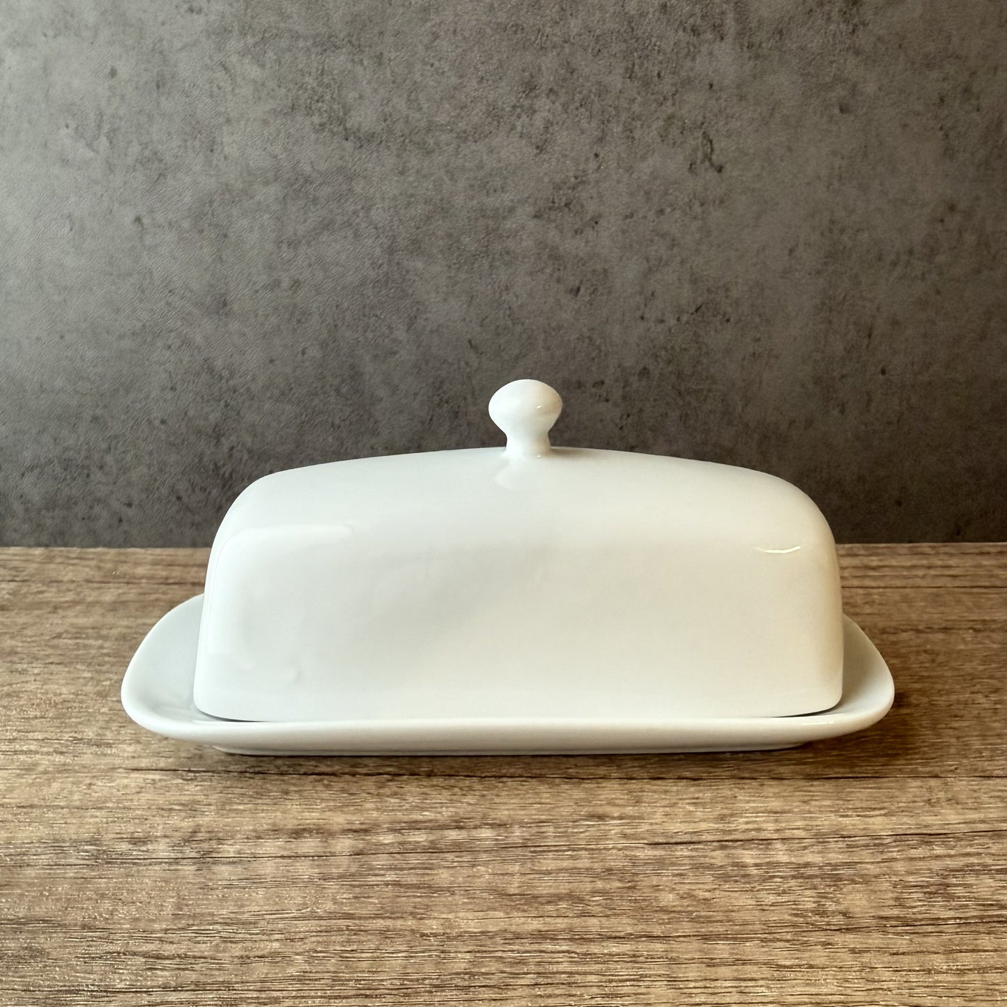 White ceramic butter dish with lid on a wooden surface against a gray wall.