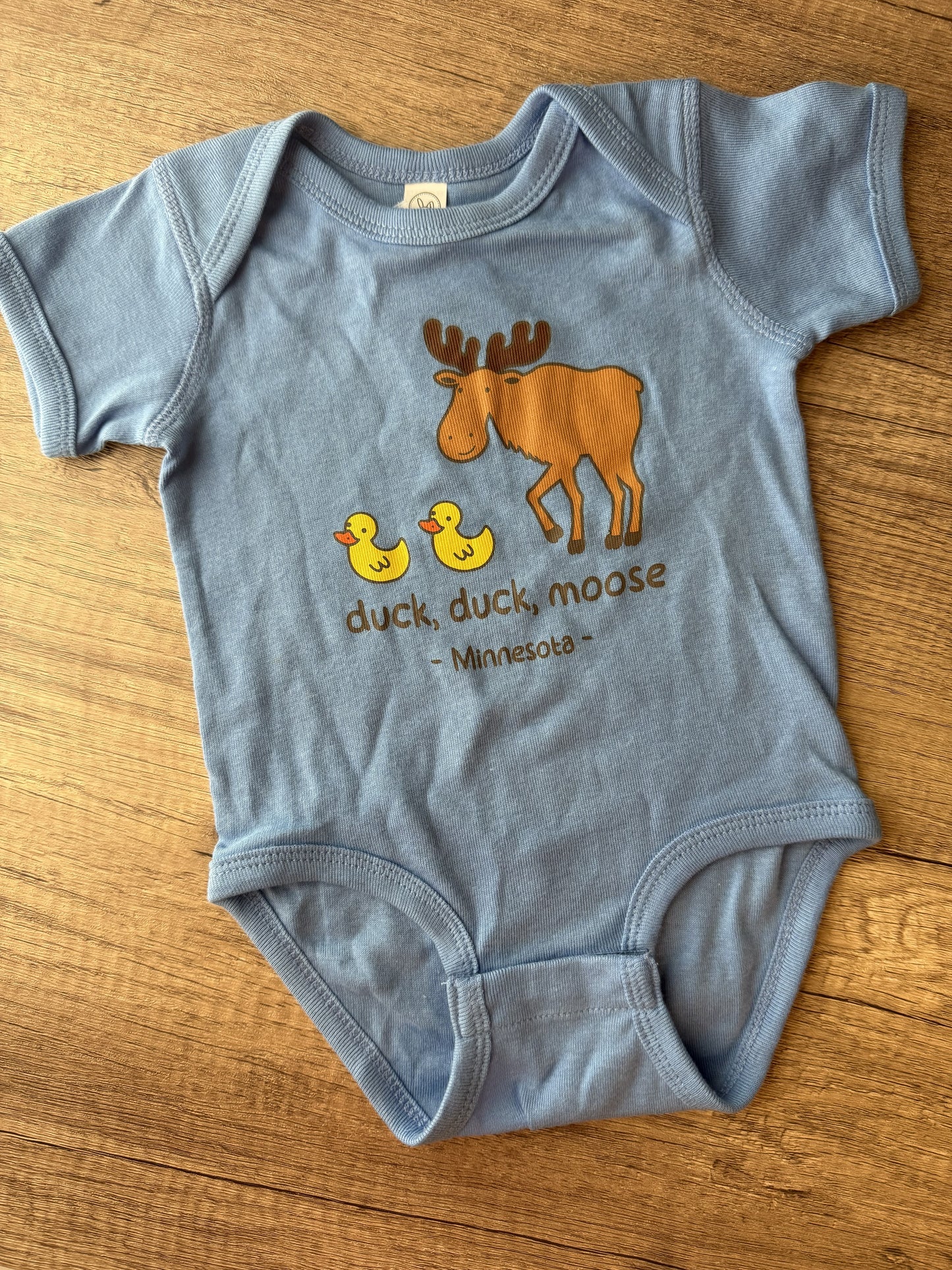 A light blue baby bodysuit laid flat on a wooden surface, featuring a graphic of a brown moose, two yellow ducks, and the text 'duck, duck, moose - Minnesota-'.