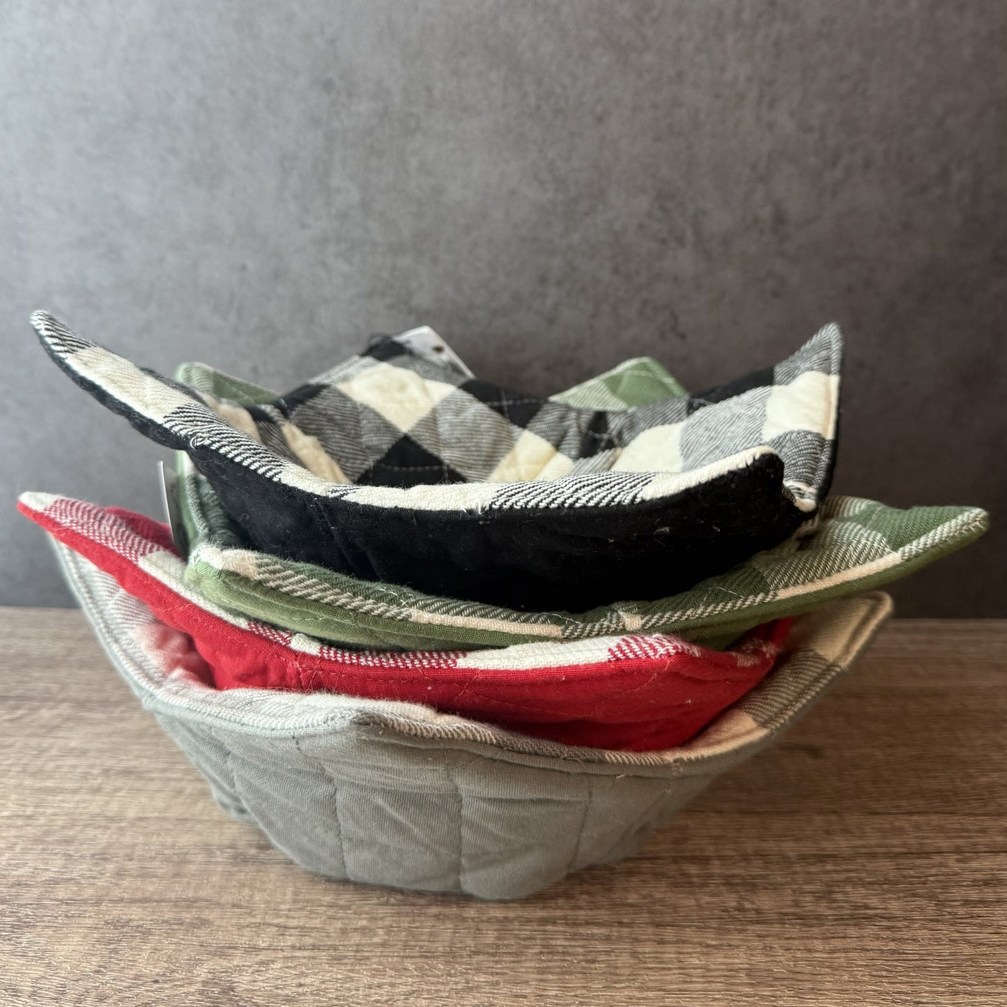 A stack of quilted fabric potholders in various patterns including plaid and solid colors, resting on a wooden surface against a gray wall.