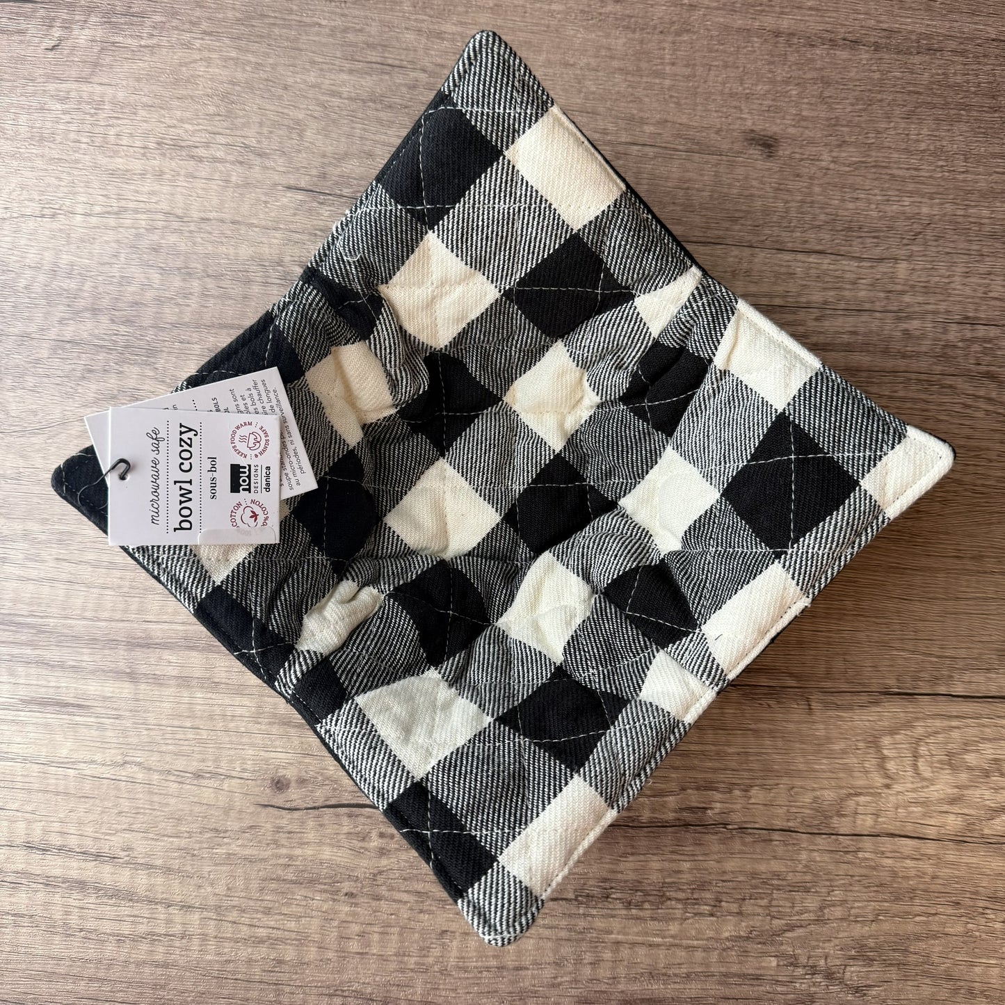 A square quilted pet bowl with a black and cream buffalo plaid pattern, shown on a wooden surface with its product tag attached.
