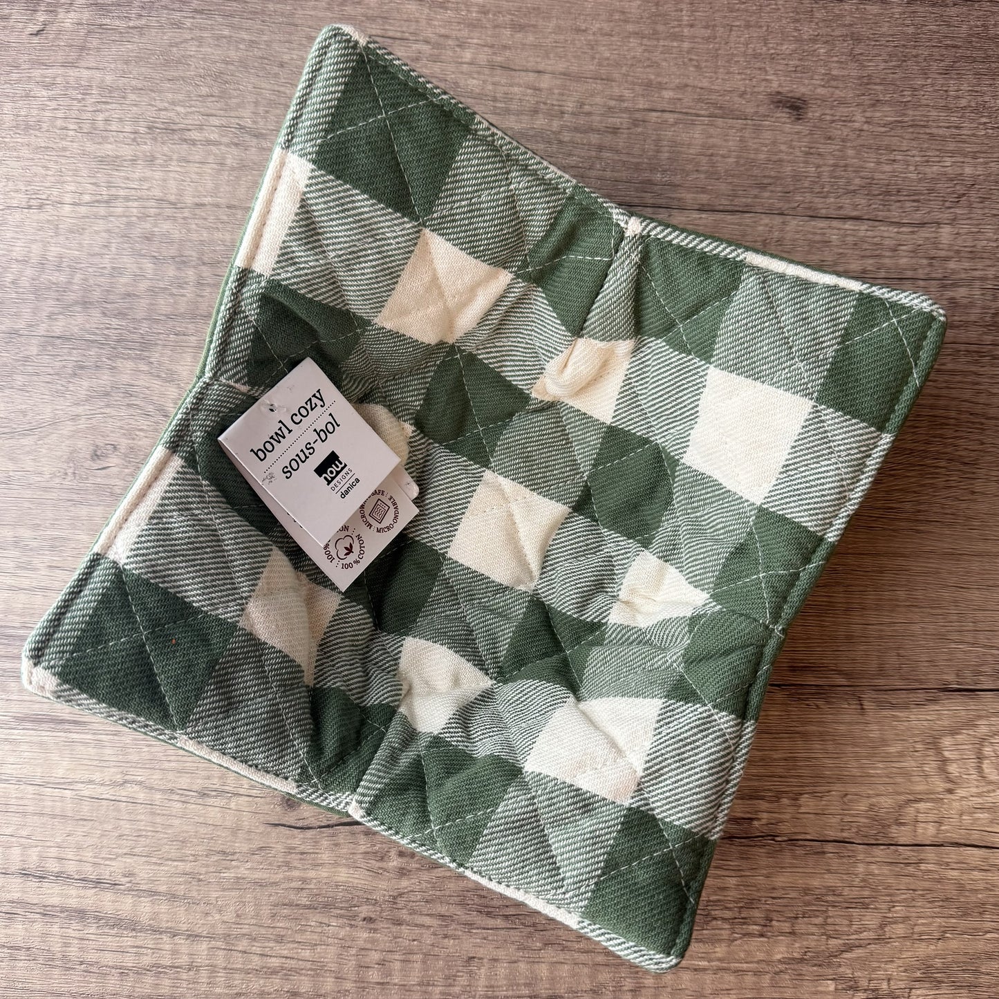 A quilted fabric potholder with a green and cream buffalo plaid pattern, featuring a sewn-in paper tag that reads 'bow tie soy-cool'. The potholder is photographed from an overhead angle on a rustic wooden surface.