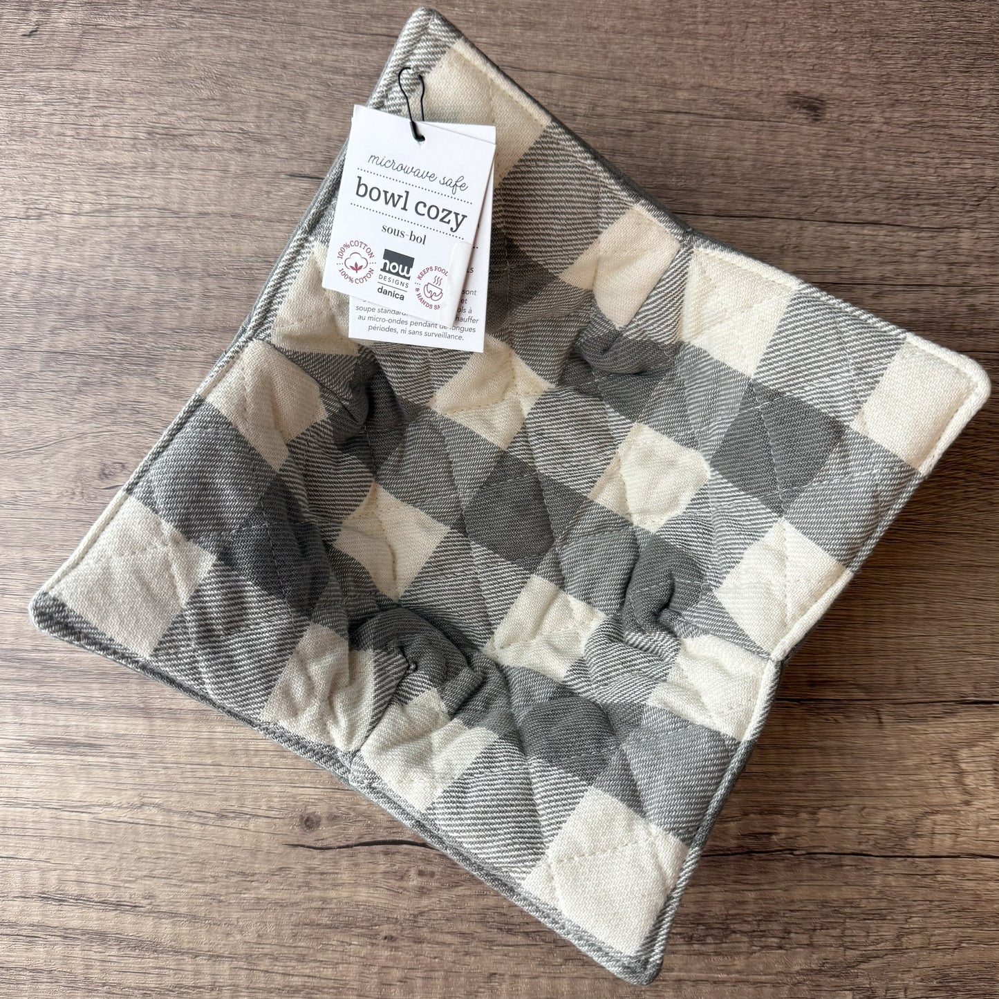 A square quilted pet bowl cozy with a gray and cream buffalo plaid pattern, shown on a wooden surface with its product tag attached.