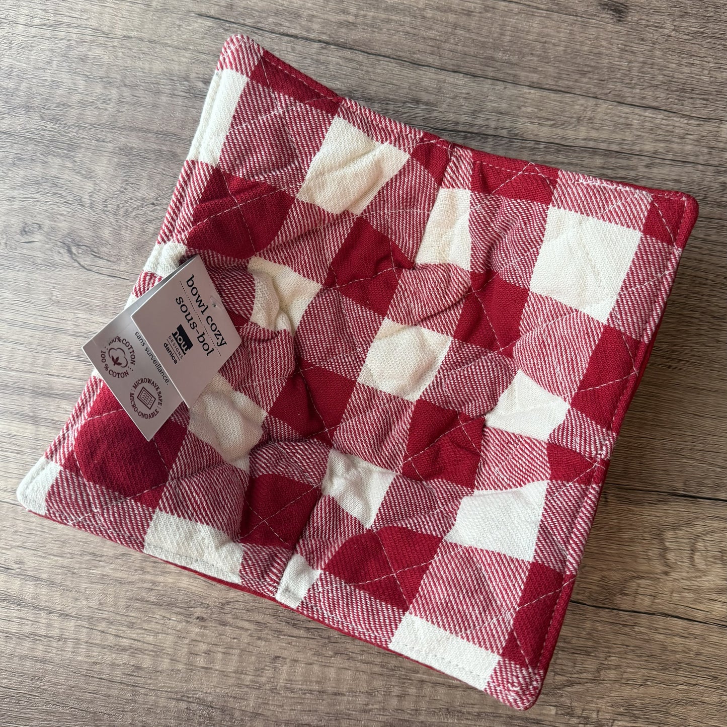 A quilted red and white gingham check counter cushion with a white paper tag attached, placed on a light brown wooden floor.